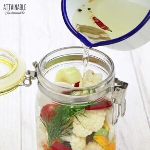 saucepan of vinegar brine being poured over a jar full of mixed vegetables.