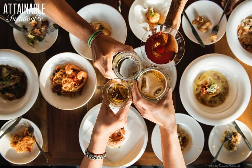 Four hands toasting with drinks from above, white plates full of food below