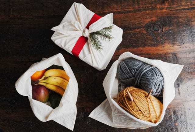 3 white bento bags on a wooden table, one christmas wrapped, one with fruit inside, one with yard skeins