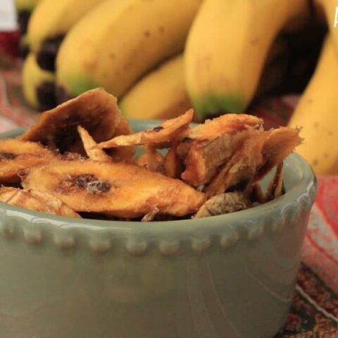 Dehydrated bananas stacked in a green bowl on a reddish striped tablecloth. Fresh bananas in the background