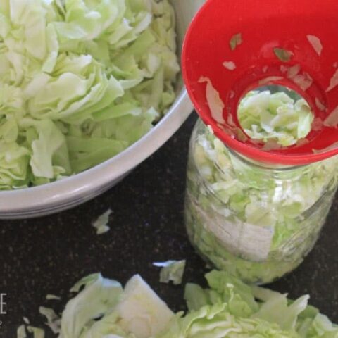 bowl of sliced green cabbage, red jar funnel with cabbage in jar