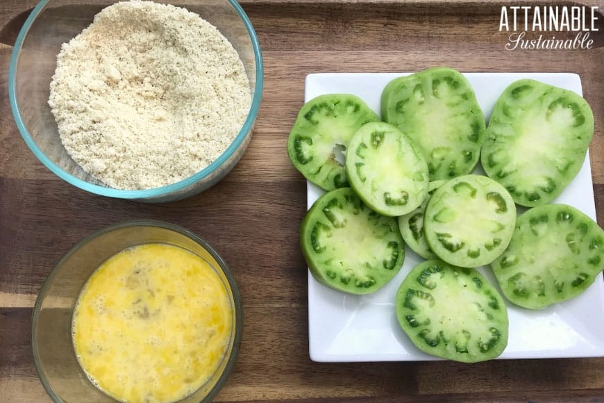 two glass bowls, one with almond flour, the other with eggs, and sliced green tomatoes on a square white plate