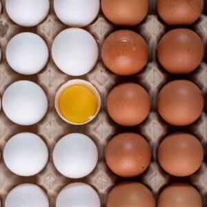 white and brown eggs in a cardboard egg carton, from above, white on left, brown on right, with one white egg cracked and the inside visible.