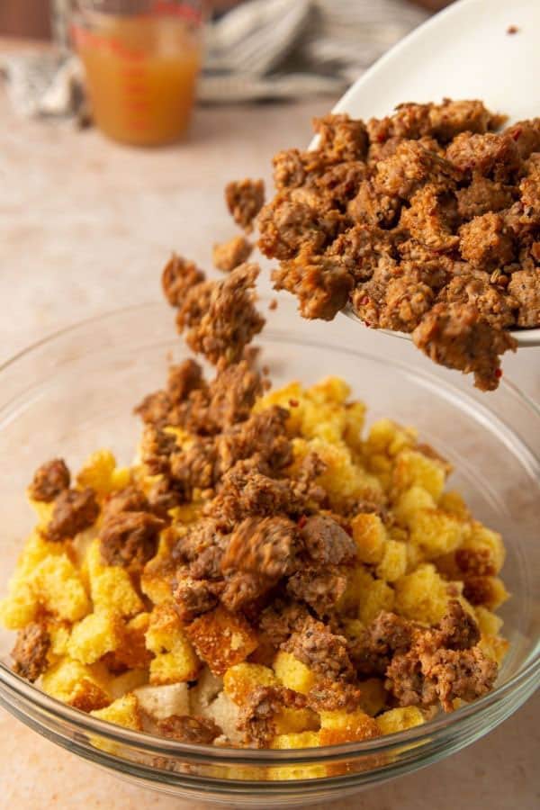 adding cooked sausage to cornbread cubes in a glass bowl