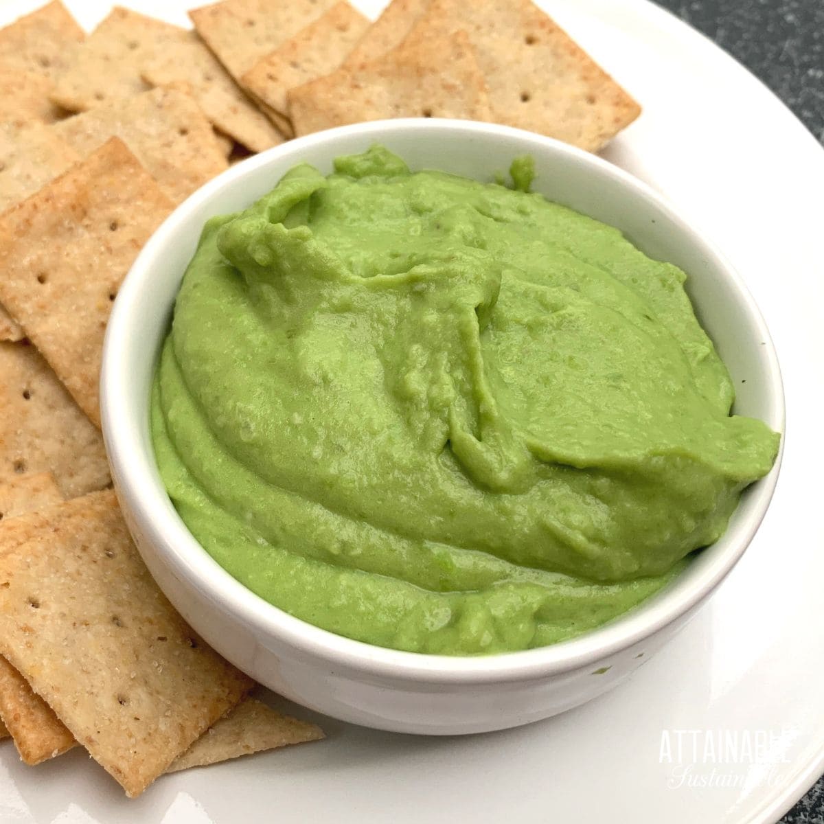 white bowl filled with bright green fava bean hummus, crackers on the side.