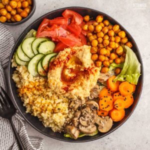 bowl from above, with cucumbers, hummus, tomatoes, carrots, grains.