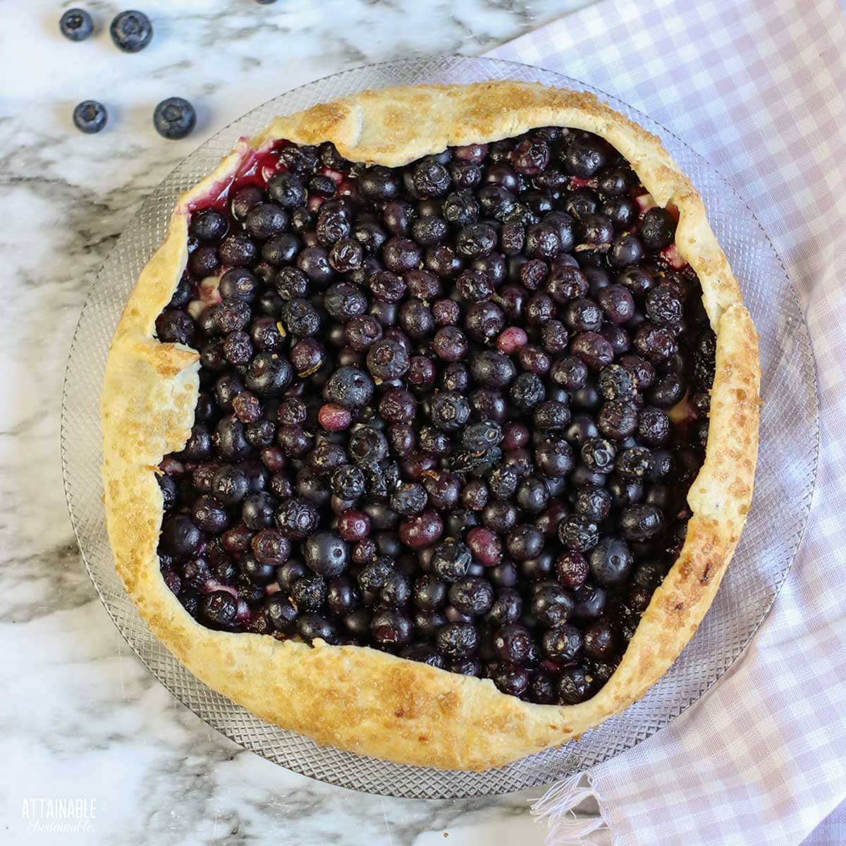 A blueberry galette on a light checkered cloth surrounded by fresh blueberries. 