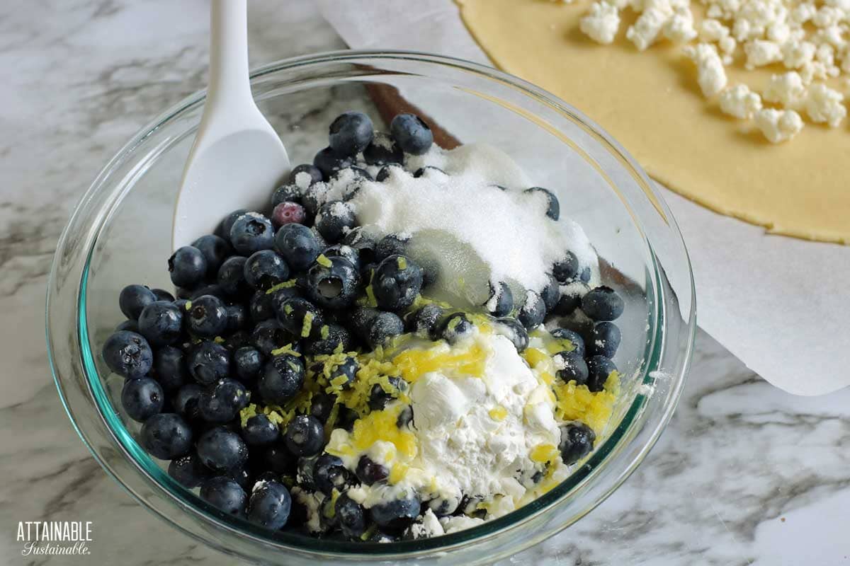 Clear glass large bowl with a white spoon mixing blueberries, lemon zest, sugar, and cornstarch together.