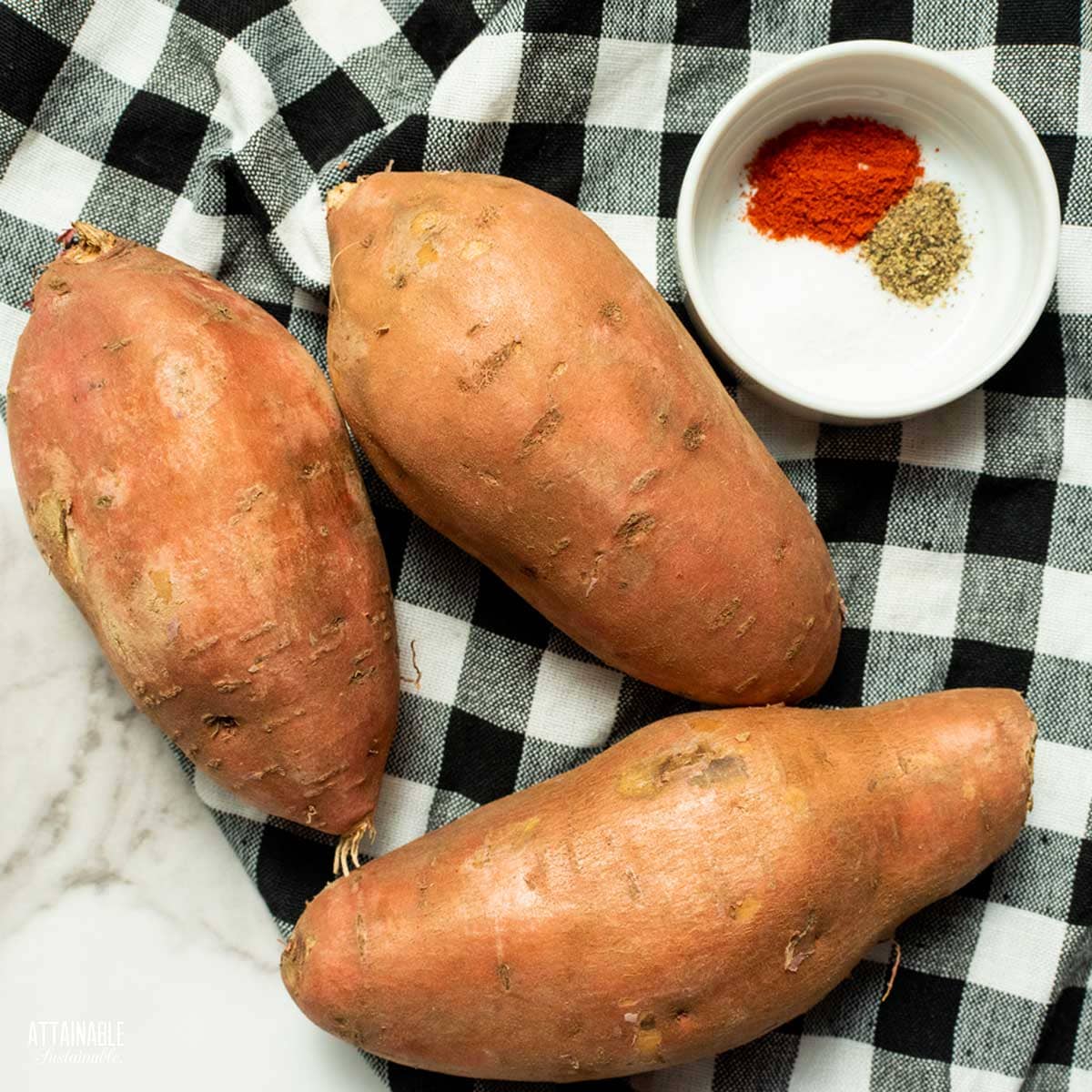 3 whole sweet potatoes with a small bowl of spices next to them, on a black and white checkered napkin. 