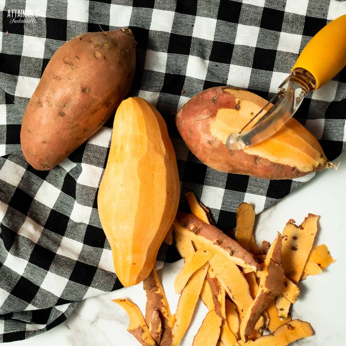 3 sweet potatoes in the process of being peeled, with the peels sititng next to them and and orange potato peeler, all on a black and white checkered cloth. 
