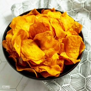 A bowl of orange crispy sweet potato chips, with a black and white patterned napkin beneath.