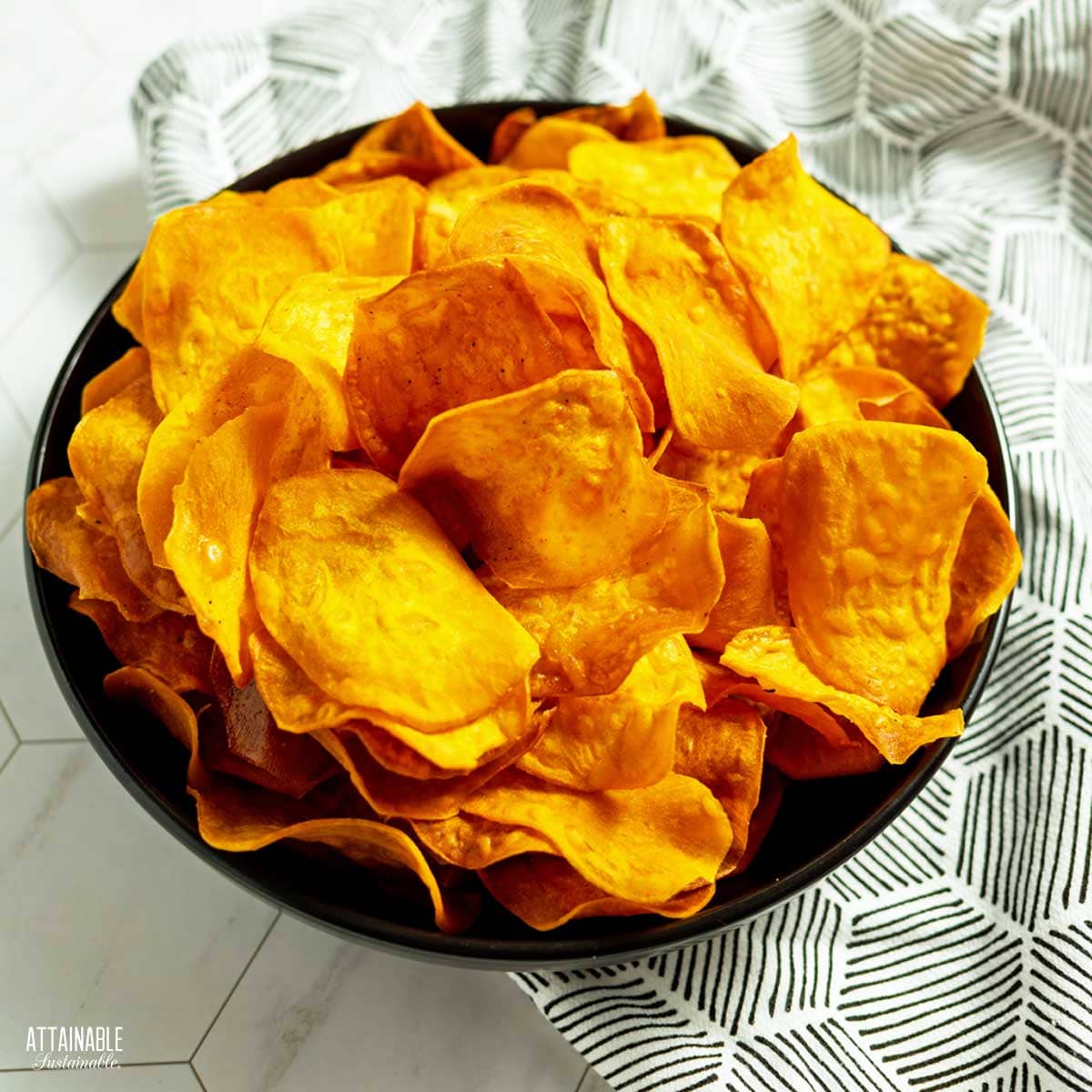 A bowl of orange crispy sweet potato chips, with a black and white patterned napkin beneath. 