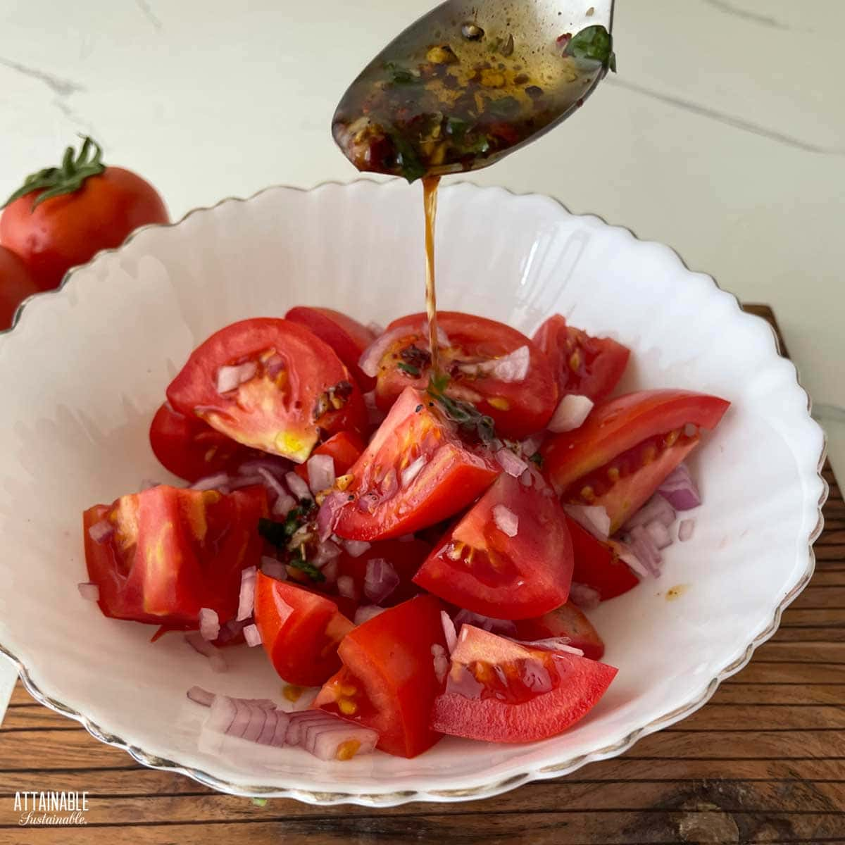 A fancy white bowl with feathered gold edges, with tomato and onion salad, and a spoon drizzling the marinade on top. 