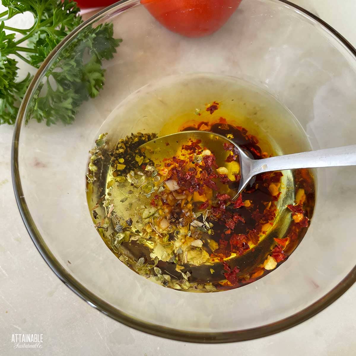 A clear bowl with the marinade ingredients being stirred with a spoon, with fresh herbs and tomatoes in the background.