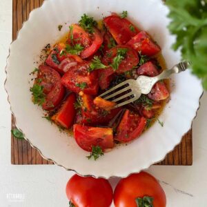 A fancy white bowl with chopped tomato and onion salad with a fork in it, surrounded by whole tomatoes.