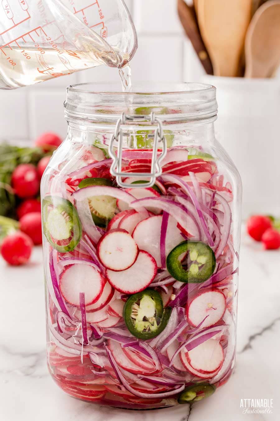 measuring cup pouring brine into swing top jar filled with sliced radishes, onions, and pepper.