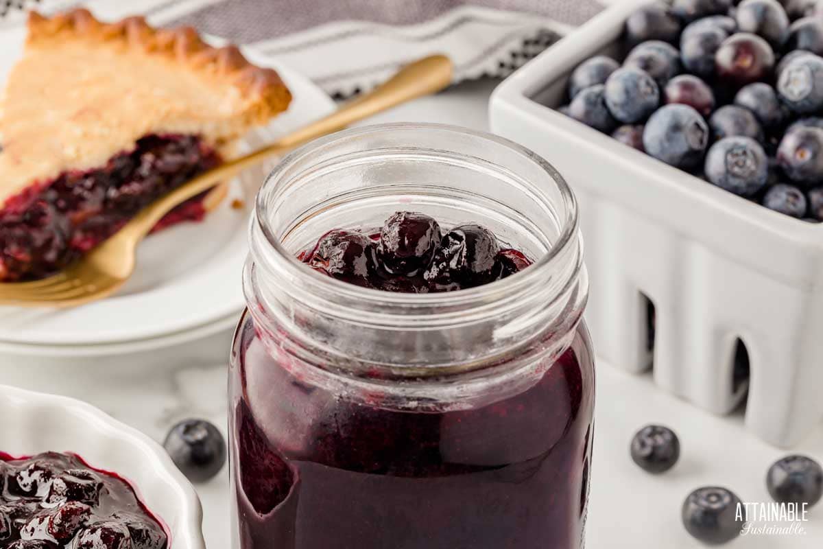 open jar of blueberry pie filling with fresh berries and a slice of pie behind.