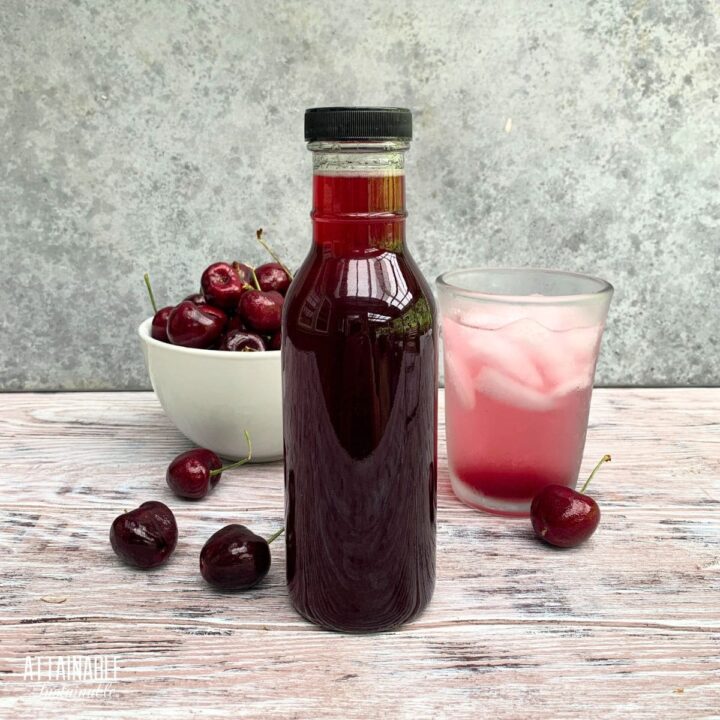 bottle of dark red cherry syrup with a bowl of cherries and an ice cold pink drink in the background.