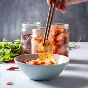 hand with chopsticks picking up fermented kimchi from a white bowl.