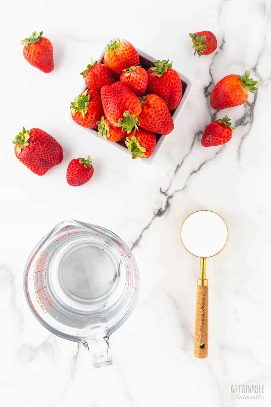 ingredients to make strawberry puree on a marble background: strawberries, sugar, and water.