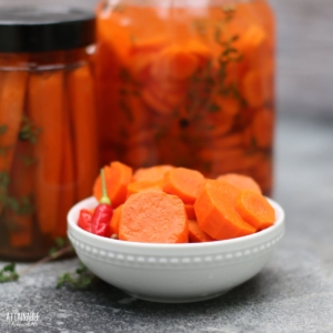 pickled carrot slices in a white bowl, with jars of pickled carrots behind.